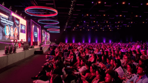 A imagem está posicionada de um ângulo alto, de forma bem ampla mostrando boa parte da plateia sentada em suas respectivas cadeiras, em frente ao palco que possui um telão enorme e aonde estão sentados 3 palestrantes.